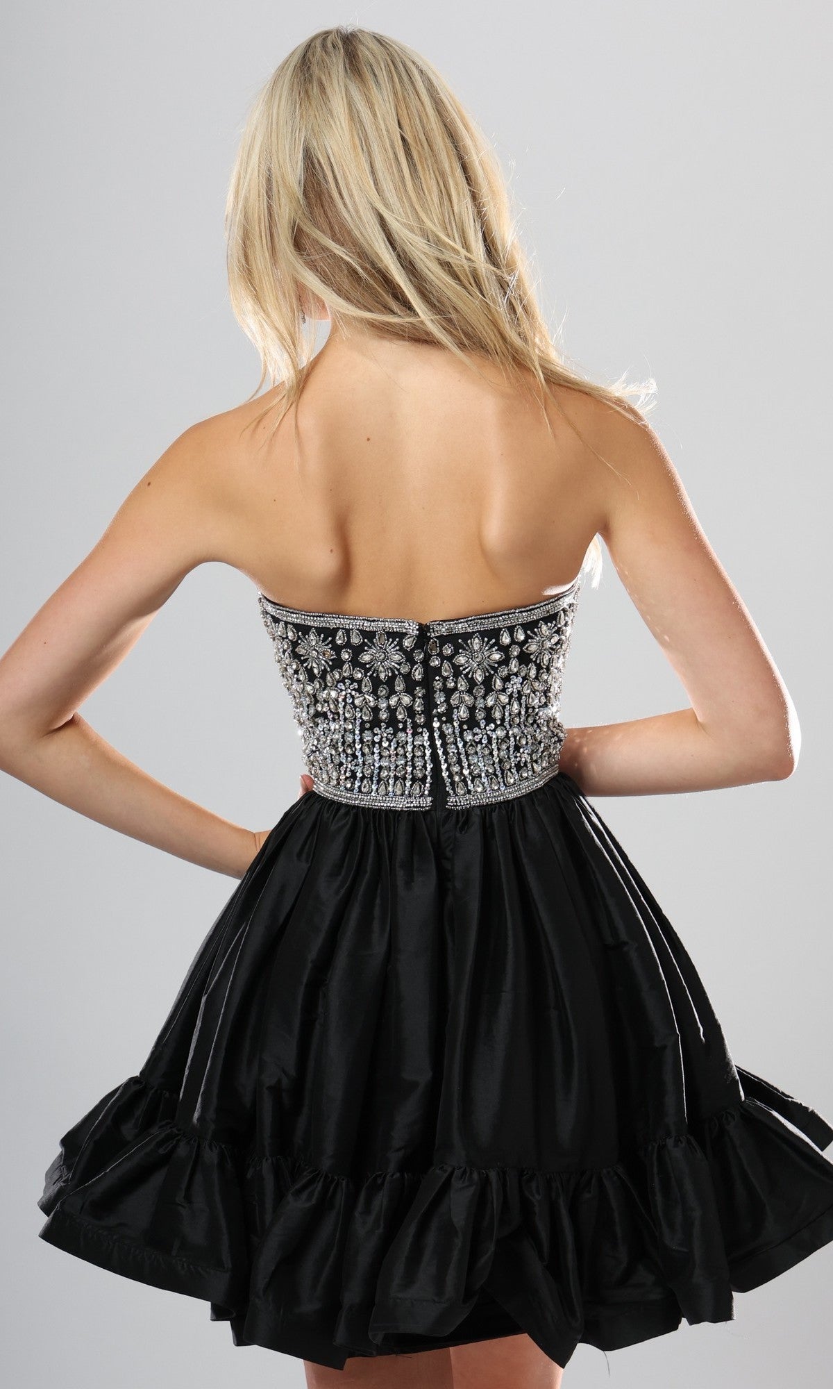 A woman with long blonde hair is shown from the back against a plain background, wearing the Jovani 52439 short homecoming dress, which features a strapless beaded bodice and a ruffled skirt.